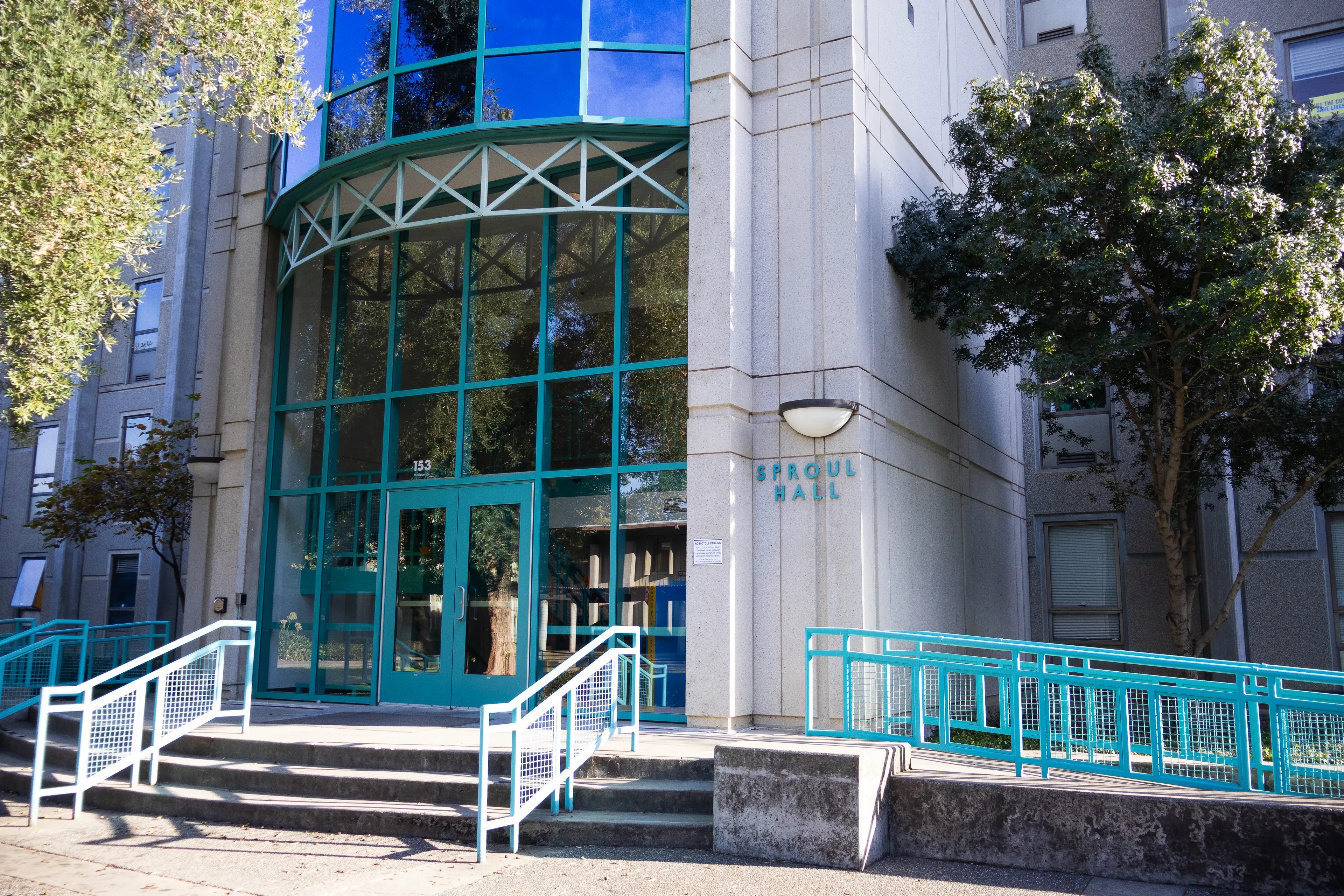 Glass-fronted office entrance with teal doors, steps and railings, flanked by trees.