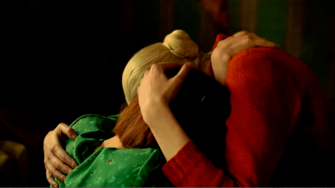 Two women, faces unseen, hold each other comfortingly. One wears green, the other red