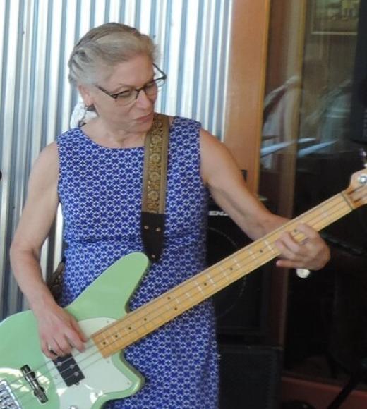 A woman with glasses wearing a blue dress with white dots playing an electric guitar.